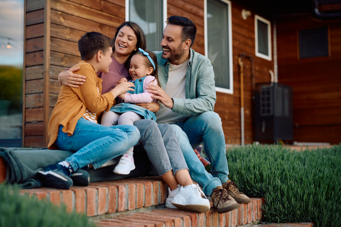A family of four — two parents and two young children — laughing together on the front steps of their home.
