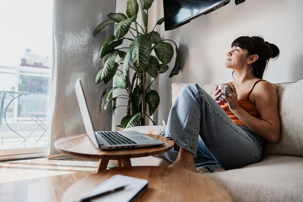 Woman sitting on her couch with a laptop and coffee, reflecting near a window, symbolizing confidence and peace of mind from accessing home equity with Unlock.