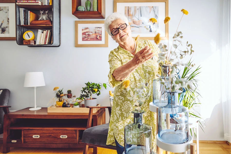 Photo of a woman arranging flowers in a vase in a sunlit room, symbolizing the confidence and comfort she has in her home.